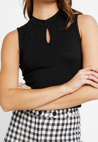 Top senza maniche nero con scollatura a chiave, abbinato a pantaloni a vita alta a quadri bianco e nero, con le braccia incrociate.