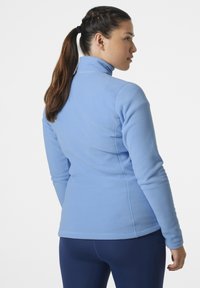 Jasnoniebieska polarowa kurtka z wysokim kołnierzem, dopasowanym krojem i płaskimi szwami. Nosi się ją na granatowych spodniach, które mają gładką teksturę.