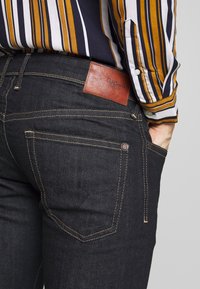 Tummansiniset farkut, joissa nahkainen merkki, näkyvät ompeleet ja kupariset niitit. Pystyraidallinen paita näkyy osittain.