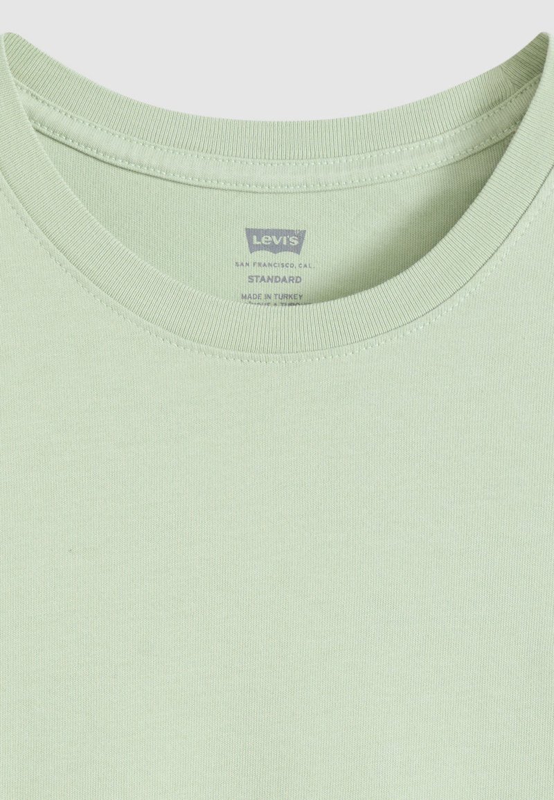 T-shirt en coton vert clair avec un col rond, une texture lisse et des bords cousus. L'étiquette indique "Levi's Standard, Fabriqué en Turquie."