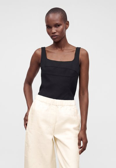 Femme portant un top noir sans manches à encolure carrée et un pantalon ample crème taille haute, debout devant un fond clair uni.