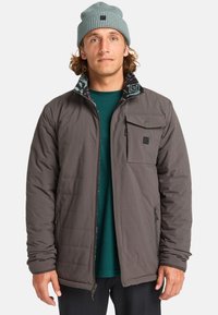 Chaqueta aislante gris con cierre zip, que presenta un cuello interior con diseño, un bolsillo en el pecho y dos bolsillos laterales con zip. Lleva puesta una parte de arriba verde.