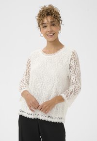 Hvid bluse i blonde med en bølget halsudskæring og trekvartlange blondeærmer. Har en bølget kant og blomsterblonde tekstur.