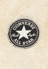 Kremowy materiał o miękkiej teksturze z czarną okrągłą łatą. Łata ma biały gwiazdkę oraz napisy "CONVERSE ALL STAR" i "Chuck Taylor."