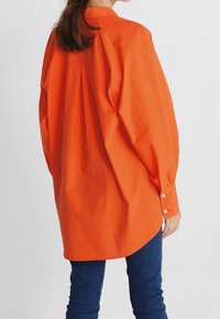 Personne portant une chemise orange à manches longues et ample, ainsi que des jeans bleu foncé, vue de dos sur un fond clair.