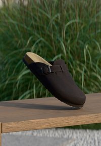 Μαύρο σουέντ παπούτσι slip-on με απαλό πάτο από φελλό και ρυθμιζόμενο λουράκι με πόρπη, τοποθετημένο πάνω σε ξύλινη επιφάνεια με φόντο φυλλώματα.