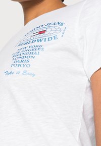 Bílé bavlněné tričko s modrým grafickým textem "TOMMY JEANS WORLDWIDE" a designem globusu, na kterém jsou vertikálně vytištěny názvy měst.