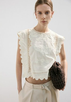 Młoda kobieta w białej, haftowanej, bezrękawnikowej bluzce i beżowych spodniach, trzymająca ciemną plecioną torebkę na tle o neutralnym kolorze.