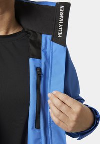 Niebieska wodoodporna kurtka z czarnymi akcentami, posiadająca kieszeń na zamek oraz miękki kołnierz. Ręka delikatnie trzymająca materiał w pobliżu zamka.