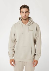 Šviesiai ruda gobtuvin�ė palaidinė su didele priekyje esančia kišene, sutraukiamomis virvutėmis ir subtiliu logotipu ant krūtinės. Minkšta medžiaga, laisvas siluetas.