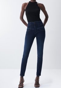 Jeans de tiro alto azul oscuro con una textura suave, dos bolsillos delanteros y cierre de botón. Combinados con un top halter negro acanalado y sandalias de tiras.