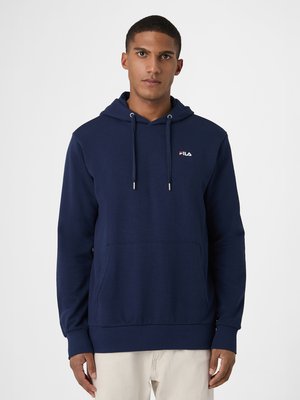 Joven con sudadera oscura FILA con bolsillo delantero y pantalones beige, de pie frente a un fondo claro y liso.