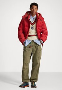 Červená prešívaná bunda, krémový pletený sveter s námorníckymi akcentmi, svetlomodrá košeľa, olivové nohavice cargo a viacfarebné turistické topánky.