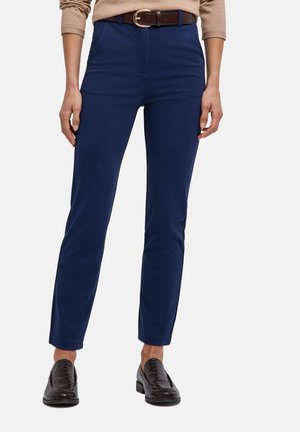 Person trägt marineblaue Slim-Fit-Hose mit braunem Gürtel, beiges langärmeliges Oberteil und dunkelbraune Loafers auf weißem Hintergrund.