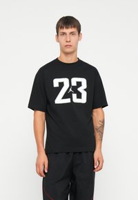 Melns īsām piedurknēm T-krekls ar lielu baltu "23" grafiku un Jumpman logo uz krūtīm; izgatavots no kokvilnas ar apaļu kakla dizainu.