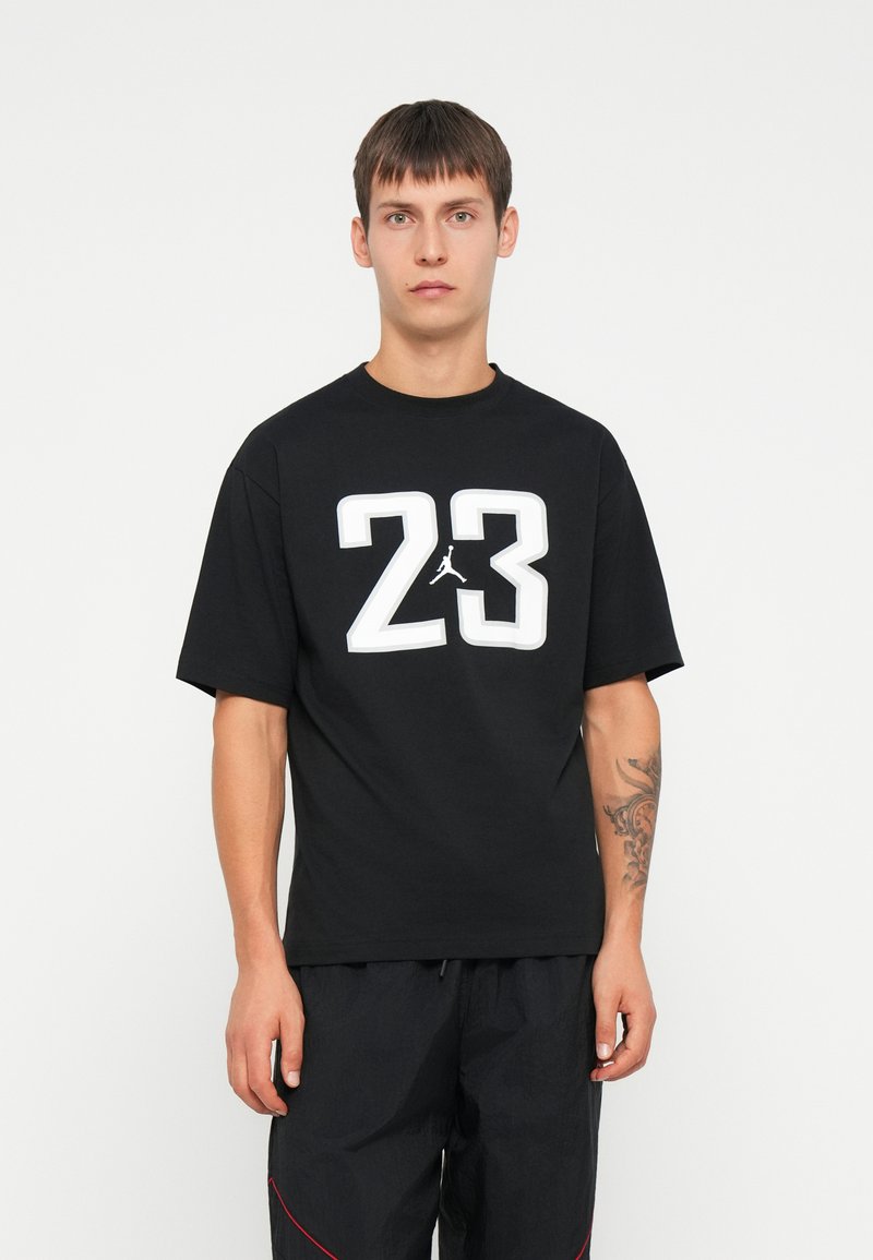 Melns īsām piedurknēm T-krekls ar lielu baltu "23" grafiku un Jumpman logo uz krūtīm; izgatavots no kokvilnas ar apaļu kakla dizainu.