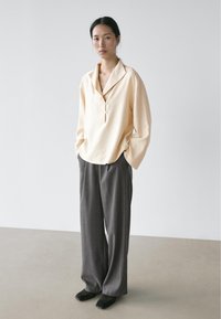 Vrouw staand met handen in haar zakken, gekleed in een crème loszittende blouse met knopen en wijde grijze broek, tegen een effen lichte achtergrond.