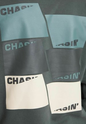 Cotton sweatshirt in dark green featuring various rectangular patches in light blue, dark gray, and cream, with "CHASIN'" printed in black.
