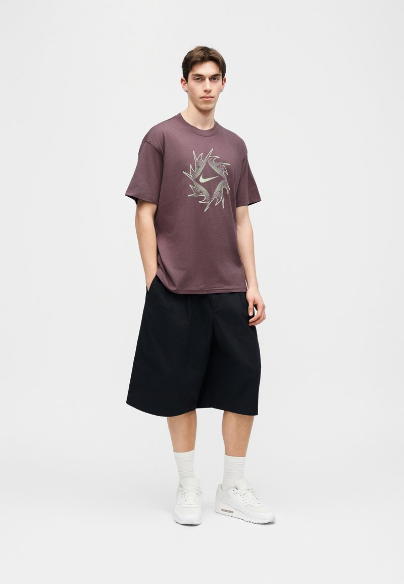 Junger Mann trägt ein lila Nike-T-Shirt mit Grafik, schwarze weite Shorts, weiße Socken und weiße Sneaker, steht mit den Händen in den Taschen.