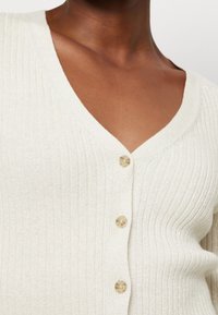 Prążkowany kardigan w jasnym kremie z dekoltem w serek, z trzema beżowymi guzikami i subtelnym metalicznym połyskiem w całym materiale.