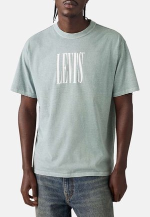 Mężczyzna w jasnizeleńskiej koszulce Levi's i niebieskich dżinsach, stoi z ramionami swobodnie opuszczonymi wzdłuż ciała.