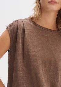 Top corto marrone a maniche corte realizzato in tessuto leggero. Presenta una vestibilità comoda, scollatura rotonda e delicate pieghe sui lati delle spalle.
