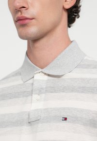 Camisa polo a rayas en gris y blanco con una textura suave, tapeta de tres botones y un pequeño logo en el pecho. Diseño clásico de cuello.