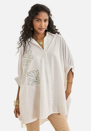 Mujer con cabello largo y rizado lleva una camisa blanca de botones extragrande con diseños florales, pantalones beige, joyas de oro y mira hacia adelante.