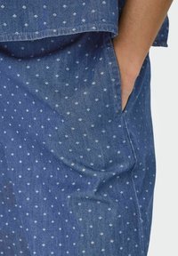 Odzież dżinsowa z niebieskiego materiału w drobne białe wzory geometryczne, boczne kieszenie i luźny krój. Teksturowany, lekki materiał.