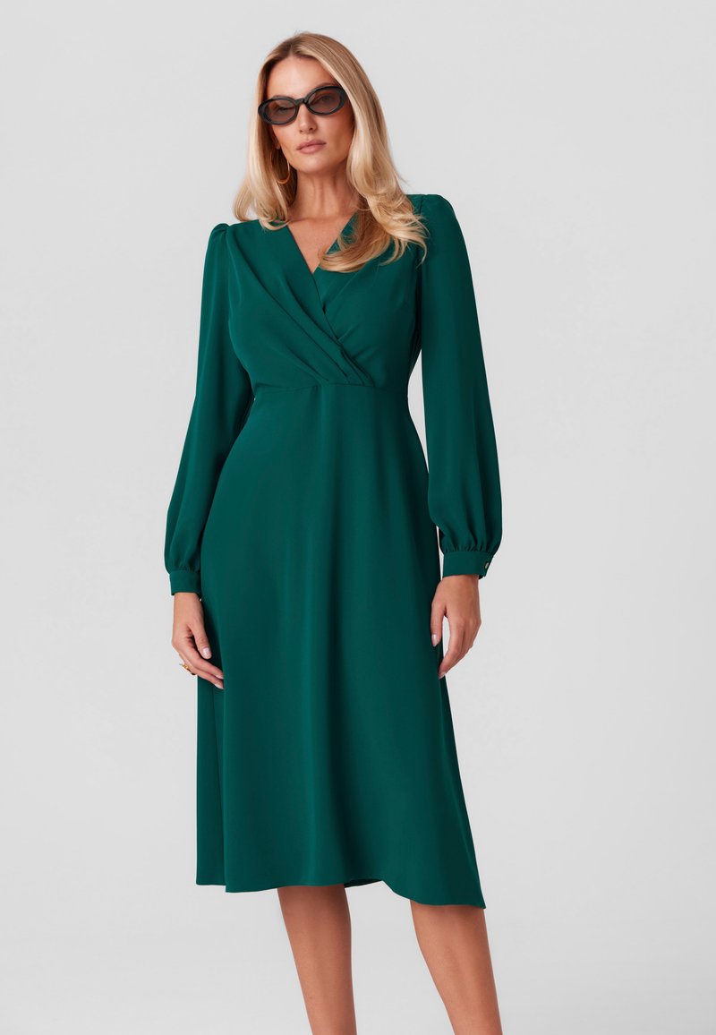 Zielona sukienka wykonana z gładkiego materiału, z kopertowym dekoltem, długimi bufiastymi rękawami i spódnicą sięgającą poniżej kolan. Bez wzorów ani akcentów.