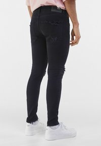 Muž v černých potrhaných skinny džínách, růžové košili, bílých ponožkách a bílých teniskách, stojící na neutrálním bílém pozadí.