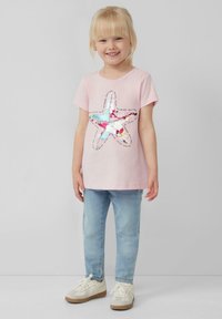 Jong blond meisje dat glimlacht, draagt een roze T-shirt met een kleurrijke paillettenster en lichtblauwe jeans, staande voor een effen achtergrond.