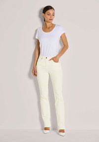 Vrouw die tegen een beige muur staat, gekleed in een witte t-shirt met korte mouwen, crèmekleurige jeans en witte puntige hakken.