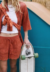 Cardigan en maille couleur rouille, haut blanc, shorts assortis avec détails brodés, tenant un skateboard vintage aux roues vertes.