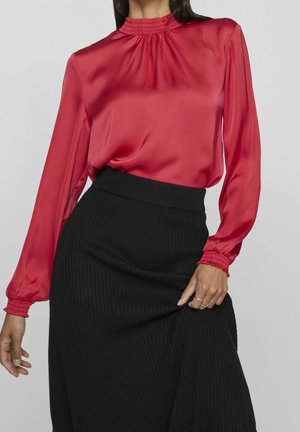 Femme portant un chemisier rouge en satin à manches longues avec col et poignets froncés, assorti à une jupe noire côtelée taille haute.