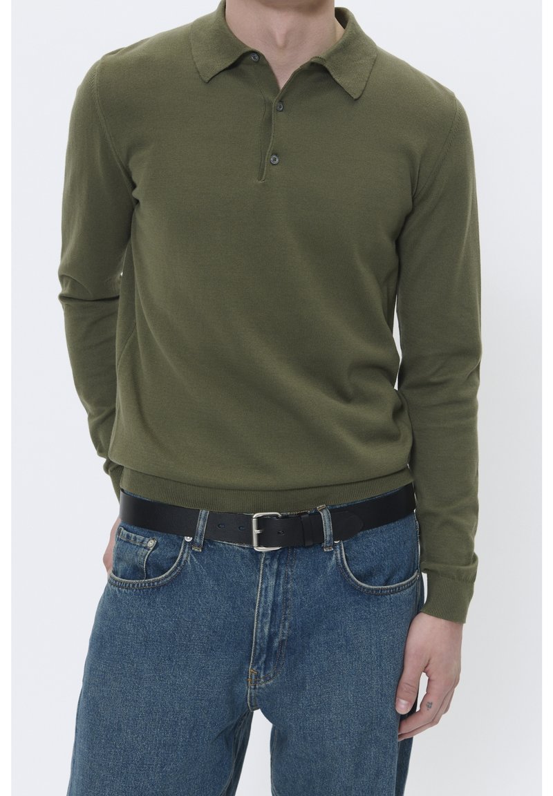 Olivgrön långärmad pikétröja med krage, treknappsstängning och ribbade ärmslut, ihop med blå denimjeans och ett svart bälte.