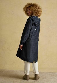 Impermeabile blu navy con motivo, cappuccio, maniche lunghe e spacchi laterali. Abbinato a pantaloni beige risvoltati e stivali marroni alla caviglia.