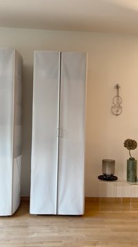 Baltos dvivėrės spintos elegantiškas dizainas ir lygus paviršius. Dešinėje pusėje ant šoninio stalelio stovi cilindro formos stikliniame vazone gėlės ir žvakė.