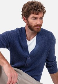 Marineblauer, gerippter Cardigan aus weichem Material, mit V-Ausschnitt, drei Knöpfen und umgeschlagenen Bündchen, kombiniert mit einem weißen T-Shirt darunter.
