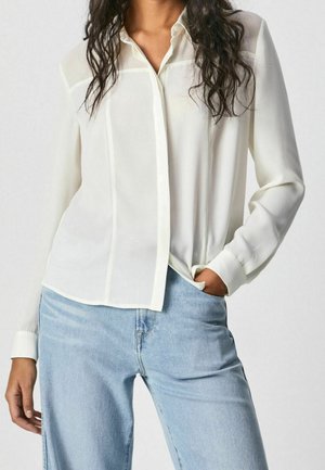 Vrouw draagt een wit overhemd met lange mouwen, lichtjes ingestopt in lichtblauwe spijkerbroek, met één hand in de zak.