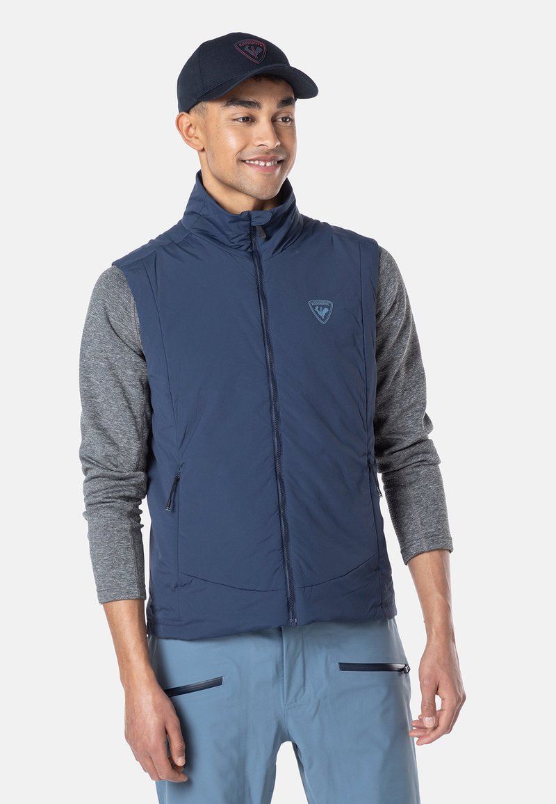 Marineblaue, isolierte Weste mit hohem Kragen, Reißverschluss vorne und Logo-Detail. Kombiniert mit einem grauen Langarmshirt und hellblauen Hosen.