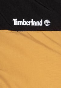 Tissu Timberland noir et jaune avec une finition texturée, présentant un logo blanc proéminent et un emblème d'arbre sur la partie supérieure noire.