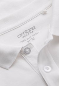 Vit bomullspolo med krage, två knappar och en texturerad tyg. Etiketten säger "ombre cotton line, 100% ren bomull, M/48."