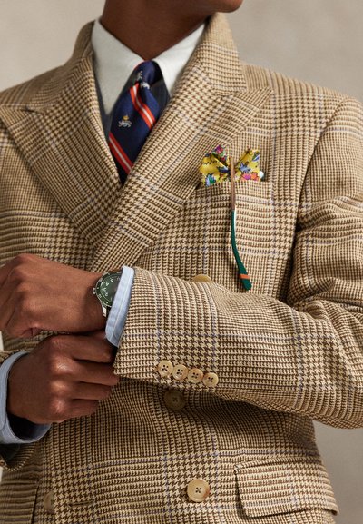 Persona che regola l'orologio al polso, indossando un blazer beige a quadri con un fazzoletto da taschino giallo a motivo floreale e una cravatta a righe blu e rosse sopra una camicia bianca.