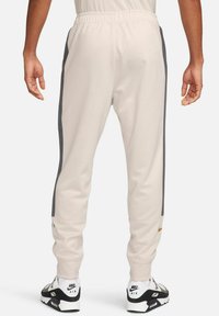 Pantalones de chándal color beige claro con franjas laterales grises, cinturilla elástica y puños acanalados. Presenta un pequeño detalle de logotipo dorado cerca del tobillo.