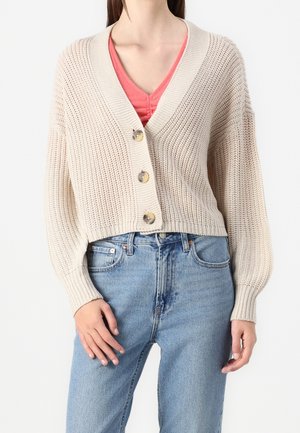 Femme portant un cardigan épais tricoté de couleur crème avec trois boutons, un haut rose à col en V et un jean taille haute bleu clair.