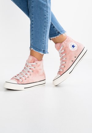 Jambes en jean bleu portant des baskets montantes Converse roses en fourrure avec des lacets blancs et des semelles en caoutchouc sur un sol blanc.