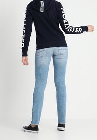 Felpa navy con testo bianco "HOLLISTER" sulle maniche, indossata con jeans slim-fit azzurri e sneaker nere. Tessuto testurizzato, vestibilità casual.