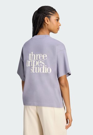 Mujer con cabello trenzado que lleva una camiseta morada holgada con el texto "three stripes studio" en la espalda y pantalones beige claros, de pie de lado.