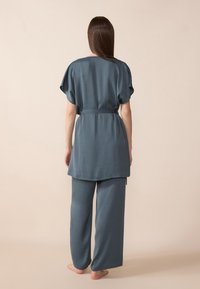 Dvojdielny outfit z tmavozelenej látky. Top s krátkymi rukávmi a viazacím opaskom, v kombinácii s voľnými, širokými nohavicami. Hladká textura.